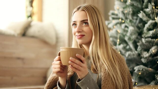 Attractive young blond woman with warm blanket in the light living room holding and drinking a cup of tea near the decorated Christmas tree. Winter holidays mood.
