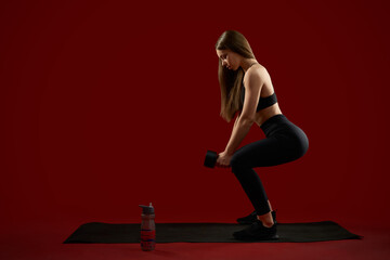 Fototapeta na wymiar Side view of woman in black training doing squatting exercises with dumbbells in red studio. Sexy woman in black have training alone workout, pump legs muscles. Concept of women in hard sport.