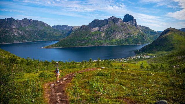 backpacker girl hiking hesten trailhead overlooking the town of fjordgard and mighty mountains in norway, senja island hiking, famous segla mountain