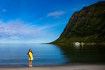 a beautiful girl in a yellow dress stares at the sea on a beach surrounded by mighty mountains on the island of senja in norway, holiday in the norwegian fjords, steinfjord
