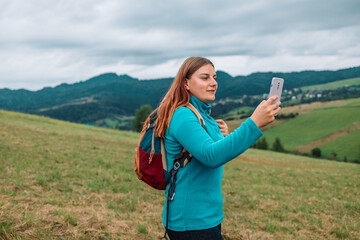 Tourist woman using smartphone and relaxing walking on background tops of mountains. Young girl traveler with mobile phone hiking summer outdoors enjoys leisure on nature. 