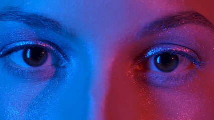 Womens eyes with shiny make up with sparkles and arrows looking straight. Look of female eyes with eyelashes and eyebrows illuminated with blue and red neon light. Macro portrait of female brown eyes.