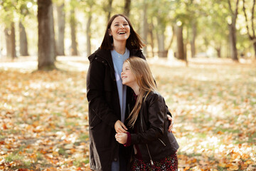 Horizontal portrait of woman joyfully pressing to chest little girl on sunny yellow forest background. Happy mother and daughter in casual black jackets walk in golden park. Family weekend, lifestyle