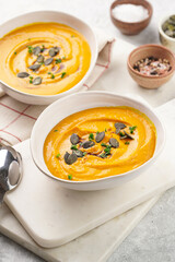 Two white soup bowls with creamy orange pumpkin soup or mash with pumpkin seeds on kitchen towel and marble board