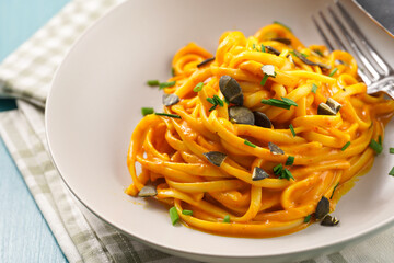 A light grey deep bowl with vegan pasta spaghetti carbonara with orange pumpkin sauce and fresh green herbs on kitchen towel on blue wooden background