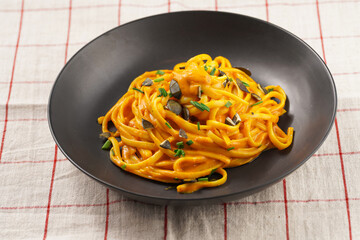 A deep black bowl with vegan pasta spaghetti carbonara with orange pumpkin sauce and fresh green herbs on checkered linen table cloth