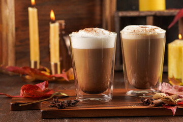 Two transparent glasses with cappuccino with black coffee, milk foam and cinnamon in autumn setting, burning candles, pumpkins, yellow and red tree leaves on wooden background