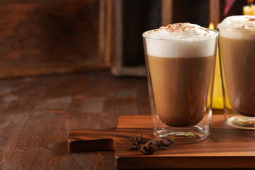 Two transparent glasses with cappuccino with black coffee, milk foam and cinnamon in autumn setting, burning candles, pumpkins, yellow and red tree leaves on wooden background