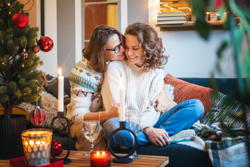 Young adult happy lesbian couple sitting on couch with christmas tree and candles at home hugging and celebrating christmas and new year eve