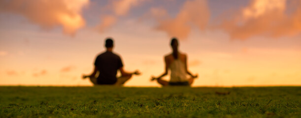 Blurred background of man woman meditating on grass facing sunset.  - obrazy, fototapety, plakaty