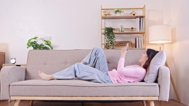 Beautiful Asian woman playing mobile game and excited about victory on sofa in living room. play games on smartphone Fun and enjoyable vacation time