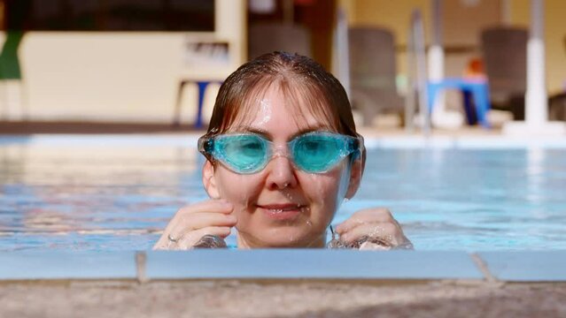 woman in swimming goggles emerges from the water of the pool. portrait of a smiling young woman doing sports
