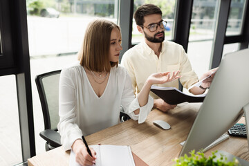 Businessman holding paper folder near new worker writing on notebook and computer in office.