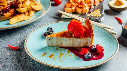 Rolgordijnen Close-up shot van een berry cheesecake op een blauw bord op een tafel met verschillende gerechten en desserts © Aleksandrs Muiznieks/Wirestock Creators