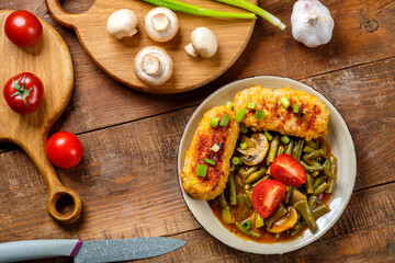 Green beans with champignons and homemade cutlets sprinkled with green onions in a plate on a wooden table next to vegetables on cutting boards and a knife.