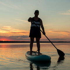 A man in shorts and a T-shirt on a SUP board against the backdrop of the sunset sky in the lake.