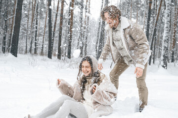 Romantic snow love story.Young couple guy girl lying,playing in snowy winter forest with trees.Walking, having fun, laughing in stylish warm clothes, fur coat,woole jacket,shawl.Date,vacation weekend