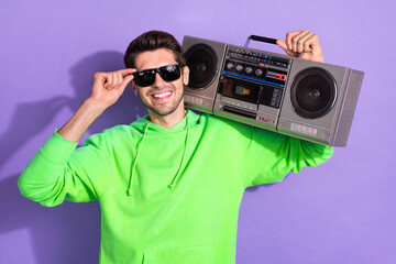 Portrait of young cheerful attractive cool student guy smiling touch specs hold boombox listen loud music disco isolated on purple color background
