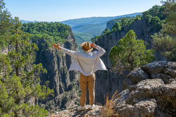 Guy in a white shirt and a brown hat, raising his hand, stands on a rock near a cliff in front of...