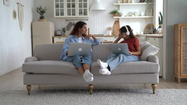 Cheerful woman asking for earphones from teenage girl sits next to on sofa with laptop on lap. Caucasian mother and daughter of school age are relaxing together using Internet and listening to music