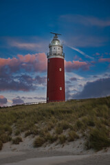 Old red lighthouse next to the dunes on Texel, Netherlands. Iconic red lighthouse in front of dark blue sky and purple clouds. Lighthouse during sunset. Typical northern sea island sunset scene. - obrazy, fototapety, plakaty