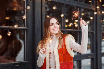 Beautiful girl in woolen pachwork vest with sparklers celebrating Christmas or New Year holiday, standing next to the garlands of light outdoors in winter. 