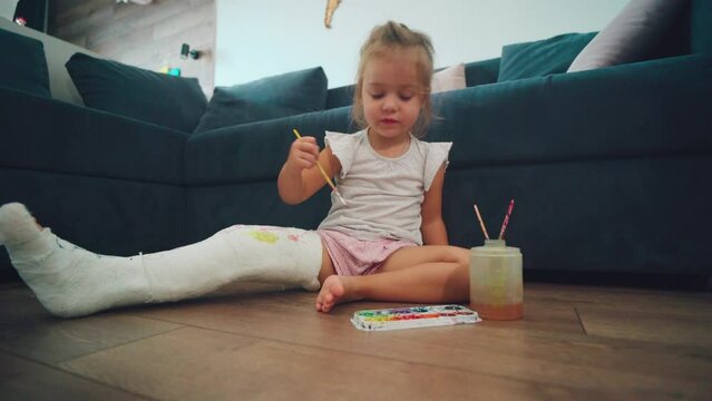 A child with a broken leg paints with watercolor paints on a plaster cast. 