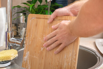 A man washes a wooden bamboo cutting board in the kitchen sink under running water. Gentle hand...