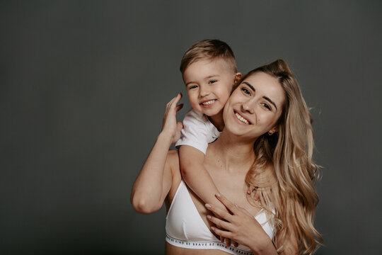 Portrait of smiling mother with curly hair and white bra posing at studio with a little son. Joyful boy embracing his mother around neck and looking together into camera