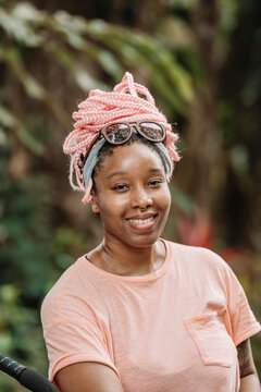 Retrato vertical de una hermosa y joven mujer afroamericana con cabello rosado muy sonriente al aire libre en un d&iacute;a de verano mirando a c&aacute;mara. 