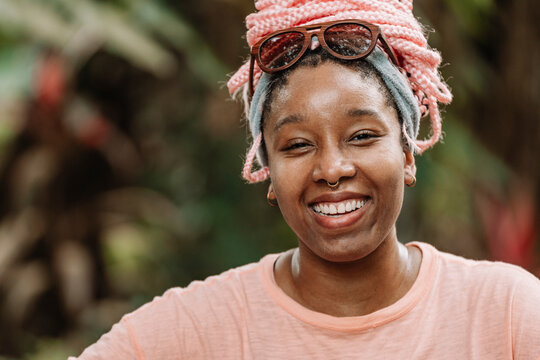 Retrato horizontal de una hermosa y joven mujer afroamericana con cabello rosado muy sonriente al aire libre en un d&iacute;a de verano mirando a c&aacute;mara. 