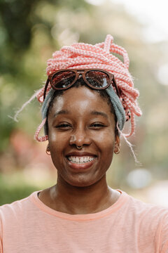 Retrato vertical de una hermosa y joven mujer afroamericana con cabello rosado muy sonriente al aire libre en un d&iacute;a de verano mirando a c&aacute;mara. 
