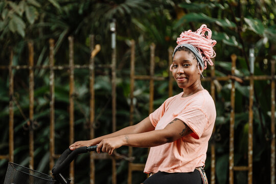 Imagen horizontal de una hermosa y sonriente mujer afroamericana con cabello rosado al aire libre con su bicicleta mirando a c&aacute;mara.