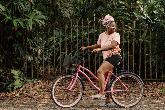 Imagen horizontal del perfil de una joven mujer afroamericana con cabello rosado en exterior dando un paseo en bicicleta en un d&iacute;a de verano. 