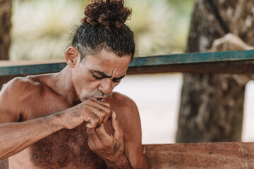Imagen horizontal de un hombre adulto de cabello rizado sin camisa en la playa encendiendo su pipa para fumar hierba.  - obrazy, fototapety, plakaty