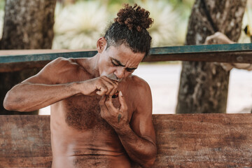 Imagen horizontal de un hombre adulto de cabello rizado sin camisa en la playa encendiendo su pipa para fumar hierba.  - obrazy, fototapety, plakaty