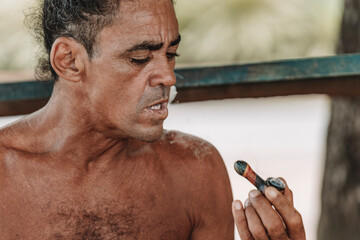 Imagen horizontal de un hombre adulto latino con cabello rizado sin camisa en la playa fumando hierba con una pipa en un día de verano.  - obrazy, fototapety, plakaty