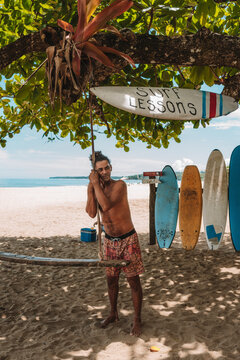 Retrato de un hombre local en el caribe de Costa Rica sin camisa recostado de un mecate que cuelga debajo de un arbol de almendro en la playa, esperando clientes en su negocio de lecciones de surf