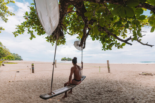 Imagen de un hombre de cabello rizado sin camisa sentado en una tabla colgando de un &aacute;rbol en una playa del caribe de Costa Rica 