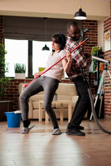 Enthusiastic multiracial couple playing guitar with vacuum cleaner and mop as finishing cleaning apartment in the city. Happy family having tidy and clean home, fun housekeeping routine.