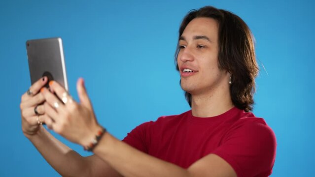Using tablet computer, happy smiling Hispanic Latino gender fluid young man 20s in red shirt talking waving in video chat call isolated on solid blue background studio portrait