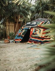 Traditional indigenous sweating hut covers by colorful Mexican blanket in tropical white sand garden with palm leaves in the foreground on a sunny day in Tulum