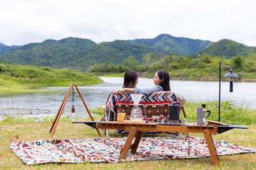 Asian woman friends drinking coffee together while having breakfast by the lake at the camp in the morning. Attractive girl enjoy outdoor lifestyle camping in forest mountain on summer travel vacation