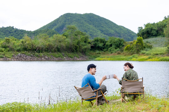 Asian man and woman friends drinking coffee together while having breakfast by the lake in the morning. Happy family enjoy outdoor lifestyle camping in forest mountain on summer travel vacation.