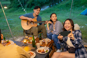 Group of Happy Asian man and woman friends enjoy and fun outdoor lifestyle using mobile phone taking selfie or video call while camping and having dinner together on summer travel vacation.