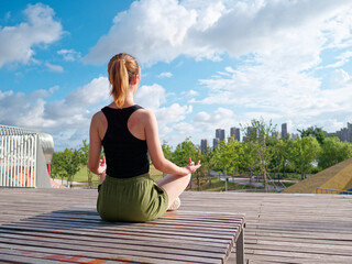 Balance with nature and the city. Rear view of young beautiful fit woman in sportswear doing yoga while sitting and meditating in city park in sunny summer day.