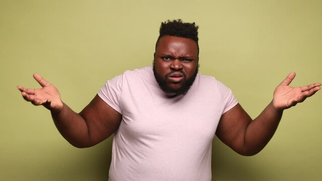 Annoyed frustrated bearded African-american man wearing pink t-shirt standing with raised hands and indignant face asking why. Indoor studio shot isolated on light green background.