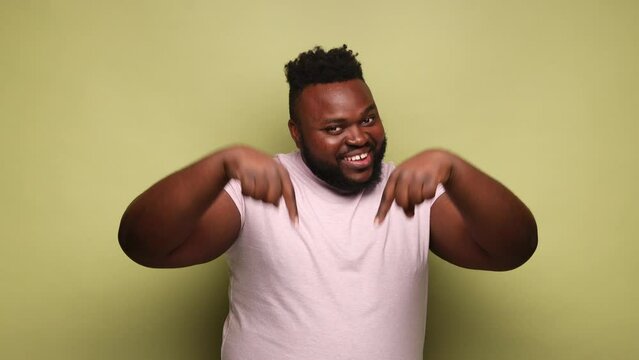Joyful African-american man wearing pink t-shirt standing looking at camera with toothy smile, pointing down, offering to subscribe. Indoor studio shot isolated on light green background.