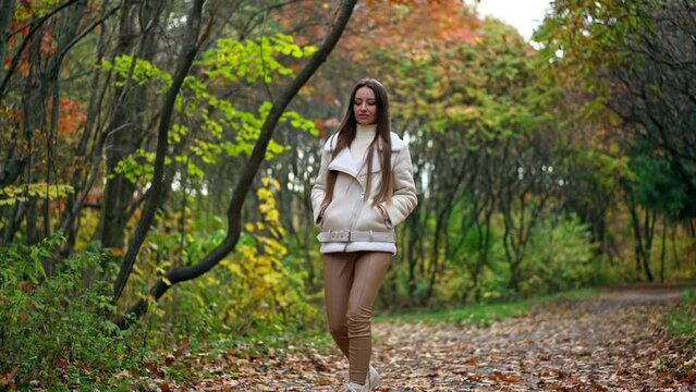 Relaxed long-haired brunette tossing fallen leaves walking by the road. Calming leisurely stride in the autumn park.