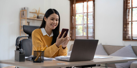 Smiling beautiful Asian businesswoman analyzing chart and graph showing changes on the market and holding smartphone at home